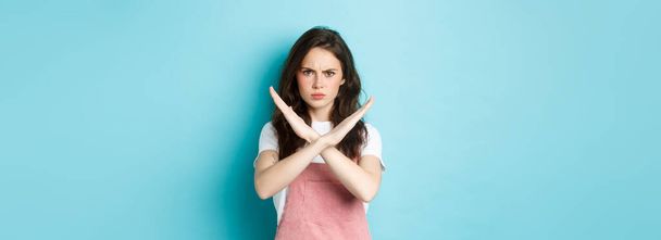 Image of young disappointed woman frowning, making cross sign and sulking, showing stop gesture, say no, forbid bad action, standing over blue background. - Photo, Image