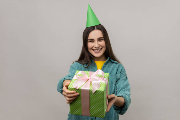 Portrait of happy woman giving green gift box to camera and smiling, sharing holiday present, congratulating on birthday, wearing casual style jacket. Indoor studio shot isolated on gray background. - Fotografie, Obrázek