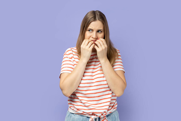 Portrait of stressed depressed blond woman wearing striped T-shirt standing looking away and biting her finger nails, having problems. Indoor studio shot isolated on purple background. - Фото, зображення