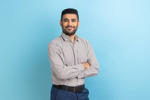 Young happy successful businessman posing with happy confident expression, looking at camera, keeping arms folded, wearing striped shirt. Indoor studio shot isolated on blue background. - Фото, изображение