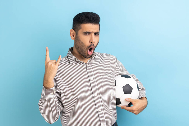 Excited businessman celebrating victory of favourite football team, holding soccer ball in hands, showing rock gesture, wearing striped shirt. Indoor studio shot isolated on blue background. - Photo, Image