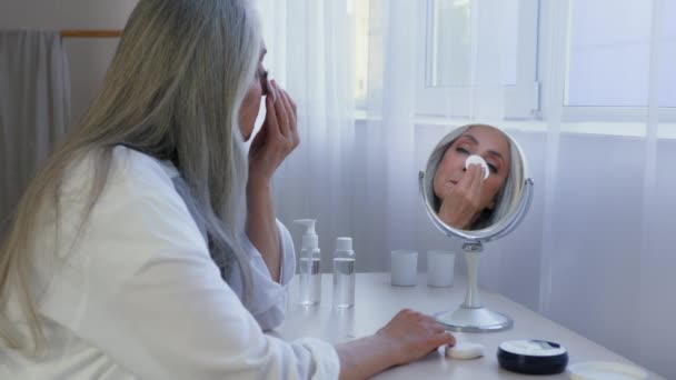 Кавказька доросла жінка середнього віку, жінка 60-х років віку, дивлячись на дзеркальні мазки, обличчям до бавовняної подушечки з тонізуючою сироваткою для лосьйонів обличчя з кремом знімає макіяж зі шкіри губки. - Кадри, відео