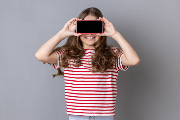 Portrait of little girl wearing striped T-shirt covering eyes with cellphone and smiling, unknown child hiding face with mobile phone, anonymous user. Indoor studio shot isolated on gray background. - Photo, Image