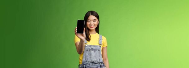Pewny siebie asertywny dobrze wyglądający kobieta azjatycki wprowadzić smartphone app, wyciągnij rękę trzymać telefon ramię, uśmiechnięty zachwycony, polecam profil społeczny, chwalić własnego bloga, zielone tło. - Zdjęcie, obraz