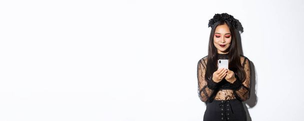Bild einer stilvollen asiatischen Frau im Halloween-Kostüm, die Nachrichten auf dem Handy checkt. Mädchen im gotischen Spitzenkleid blickt auf Smartphone, steht vor weißem Hintergrund. - Foto, Bild