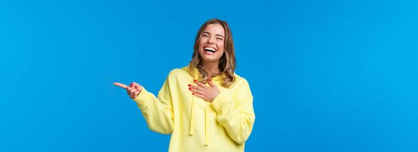 Waist-up portrait of laughing girl having fun, discuss hilarious stories with friends, chuckle touch chest as giggle carefree, look camera and smile as pointing finger right, blue background. - Photo, image