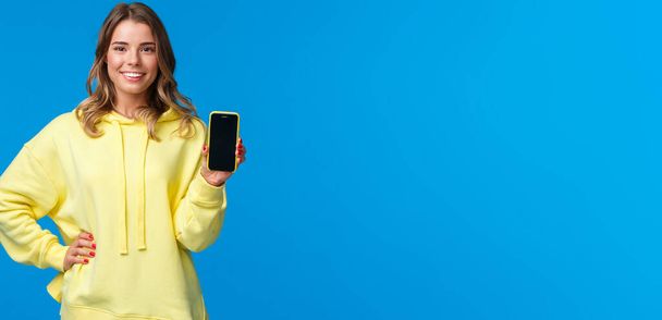 Waist-up πορτρέτο όμορφη ξανθιά κοπέλα σε κίτρινο hoodie προωθήσει την εφαρμογή smartphone, δείχνουν οθόνη του κινητού τηλεφώνου και χαμογελαστή κάμερα ως συνιστούμε εγγραφείτε ή να κατεβάσετε app. - Φωτογραφία, εικόνα