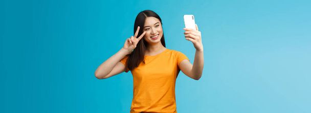 Amistoso alegre bonita mujer asiática morena corto corte de pelo tomando selfie, sonriendo alegremente mantener el teléfono inteligente mostrar paz, signo de victoria en la pantalla del teléfono cámara frontal, hablando video-llamada familiar. - Foto, imagen