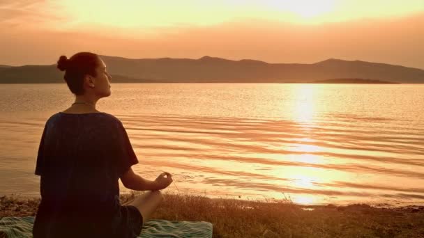 Meditating By An Ocean toont een jonge dame in silhouet die yoga doet op een houten steiger. De zon gaat onder en de omgeving baadt in een fel oranje licht. De zon zelf wordt helder weerspiegeld in het kalme rustige water.  - Video