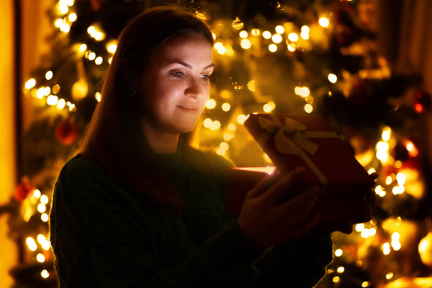 Happy beautiful woman in front of Christmas tree opening gift box and looking at the magic light inside the box. Marry Christmas and Happy New Year concept - Foto, immagini