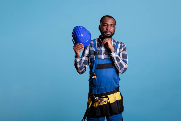 Construction worker analyzing best option to realize new renovation project. African American builder wearing work uniform and belt with tools against blue background, studio shot. - Foto, afbeelding