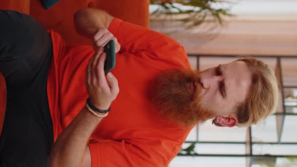 悲しいひげを生やした男は、スマートフォンのタイピングブラウジングを使用して、突然の宝くじの結果、悪いニュース、財産の損失、失敗、期限に驚いて失う。オレンジのソファの上の部屋にいる若い男。縦画面表示 - 映像、動画