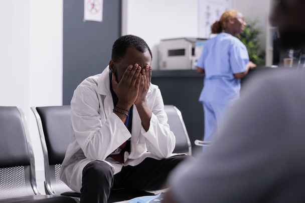 Δυσαρεστημένος δυστυχισμένος γιατρός που κάθεται στο λόμπι της κλινικής, αισθάνεται καταπονημένος και αγχωμένος στην αίθουσα αναμονής του νοσοκομείου. Κουρασμένος απογοητευμένος γιατρός να εξαντληθεί σχετικά με το φόρτο εργασίας και τις επισκέψεις ελέγχου. - Φωτογραφία, εικόνα