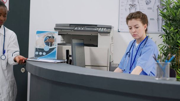 Medical assistant working at hospital reception desk in facility, helping people with healthcare appointments and checkup visits. Female nurse using report papers for consultation at health center. - Foto, immagini