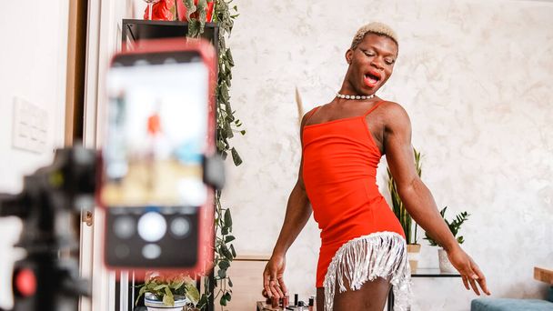 Low angle of African American man in stylish red dress dancing in front of smartphone while recording video for blog in daytime at home - Фото, изображение