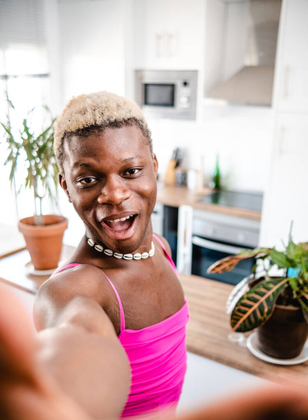 Positieve jonge Afro-Amerikaanse transseksuele man met kort geverfd haar in vrouwelijke jurk glimlachen en kijken naar de camera terwijl het nemen van selfie in de moderne keuken - Foto, afbeelding
