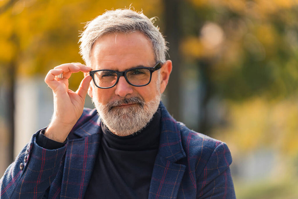 Szemüveg kereskedelmi koncepció. Kültéri közelkép egy jóképű, elegáns, 60-as éveiben járó, szürke arcszőrzetű fehér férfiról, aki fél kézzel hozzáér a fekete keretes szemüvegéhez. Meleg őszi nap süt részben - Fotó, kép