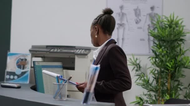 Portré afro-amerikai recepciós ellenőrzi az orvosi jelentéseket papírok hirdetés számítógép, szűrőlapok t, hogy az egészségügyi találkozók biztosítási támogatás. Regisztrációs munka a pultnál. - Felvétel, videó