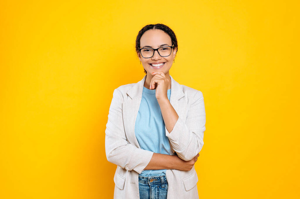 Charming beautiful positive young brunette brazilian or hispanic woman with glasses, stylishly dressed, company employee, posing while standing on isolated orange background, looks at camera, smiling - Photo, Image