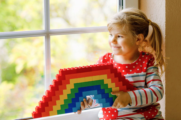 Cute little toddler girl by window create rainbow with colorful plastic blocks during pandemic coronavirus quarantine. Children made and paint rainbows around the world as sign - Photo, Image