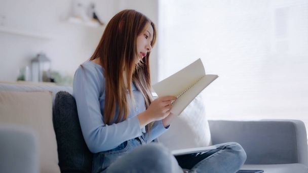 Jeune femme asiatique lisant un livre assis sur le canapé à la maison - Photo, image