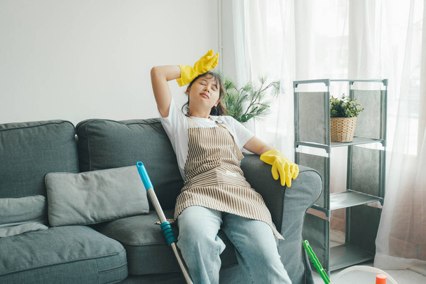 Tired and exhausted woman having rest on sofa after cleaning home or mopping floor in living room at home. Woman taking rest after cleaning home for long time. Housekeeping concept. - 写真・画像