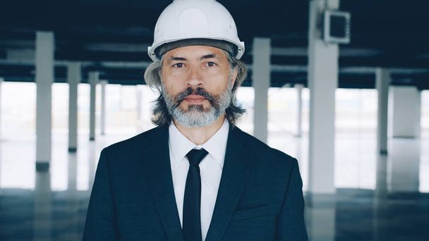 Portrait of experienced male architect standing inside new modern building wearing safety helmet and suit. Commercial property and architecture concept. - Fotó, kép