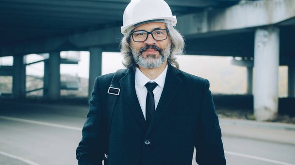 portrait of building project investor standing outdoors wearing suit and helmet and looking at camera with construction area in background - Photo, Image