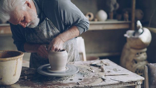 Experienced ceramist grey-haired bearded man is smoothing molded ceramic pot with wet sponge. Spinning throwing wheel, muddy work table and handmade clayware are visible. - Foto, Imagen