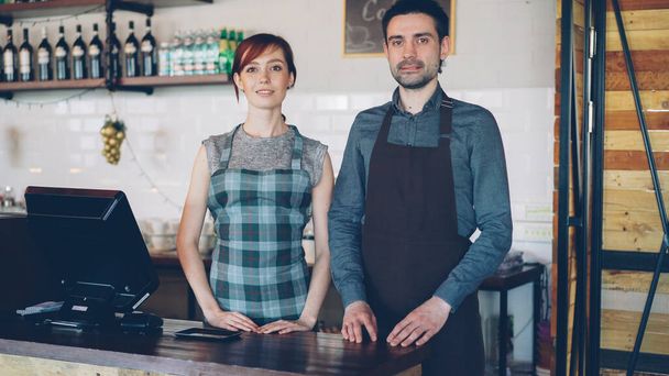 Portrait of two confident waiters in aprons standing at cashiers desk in new coffee-house and smiling. Successful business, happy people and food service concept. - Foto, Bild