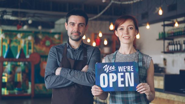 Happy attractive people cafe workers in aprons are holding yes we are open sign and smiling while standing inside coffee shop. Opening new business and start-up concept. - Φωτογραφία, εικόνα
