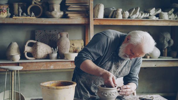 Experienced craftsman bearded man is making ear for ceramic bowl and fixing it on clayware while working in cozy workplace. Pottery, profession and hobby concept. - Foto, immagini