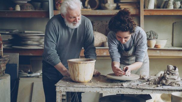 Caring grandfather experienced potter is teaching yong girl how to work with clay on potters wheel at workshop. Pottery, family hobby and handicraft concept. - Φωτογραφία, εικόνα