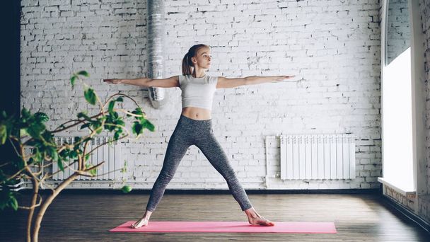 Young slim blond girl is doing yoga complex in nice studio with white walls. She is starting with warrior poses, then bending forward and backward and finishing with namaste. - Foto, imagen