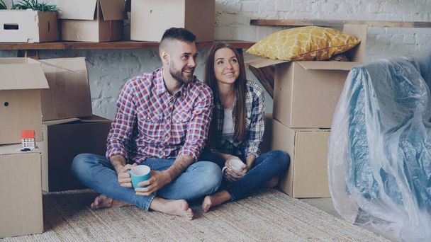 Happy couple is sitting on floor in new house and having conversation discussing interior of their new home holding mugs. Carton boxes around them are in background. - Photo, image