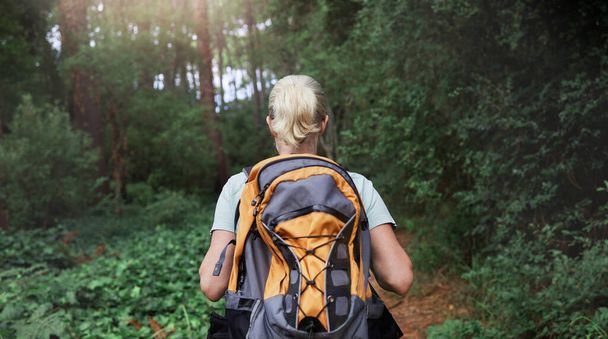 Rear view of a woman wearing a backpack hiking through a forest in nature alone. Female walking on a trail in the outdoors. - Φωτογραφία, εικόνα