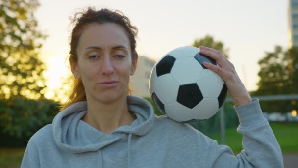 A nő, aki focilabdát tart a kamerába néz és mosolyog a napfényben. Egy női focista portréja a parkban naplementekor. Naplemente háttér. Kiváló minőségű 4k felvételek - Felvétel, videó