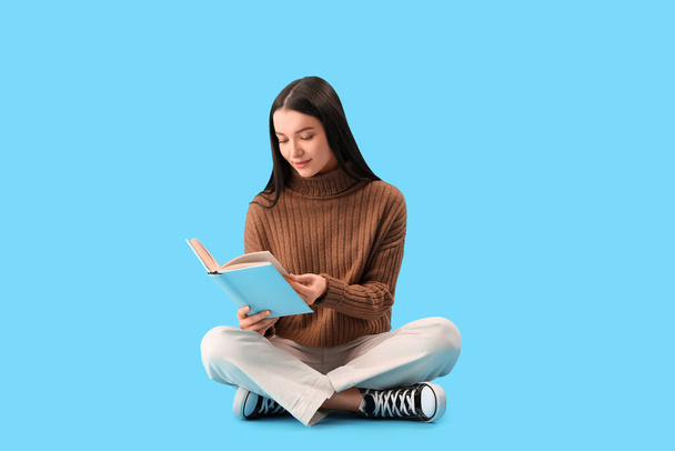 Junge Frau im braunen Pullover liest Buch auf blauem Hintergrund - Foto, Bild