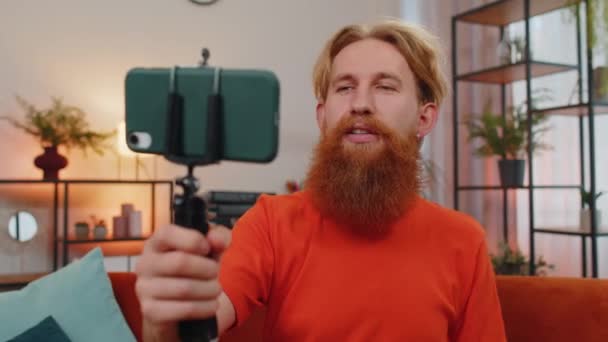 Portré kaukázusi szakállas férfi blogger csinál szelfi okostelefon állvány, kommunikáló videohívás online előfizetők. Fiatal srác a lakásban a szobában ül a narancssárga kanapén. Életmód - Felvétel, videó