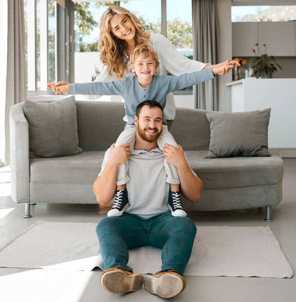 Portrait of a happy family. Parents and son at home. Adorable caucasian boy smiling and sitting on his fathers shoulders with arms outstretched. Young husband enjoying time with his wife and son. - Photo, Image