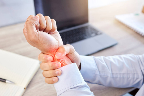 Closeup of hand and wrist pain highlighted in red. A businessman holding his arm with an injury. Male employee working at his desk in a modern office and suffering from a medical problem. - Photo, Image