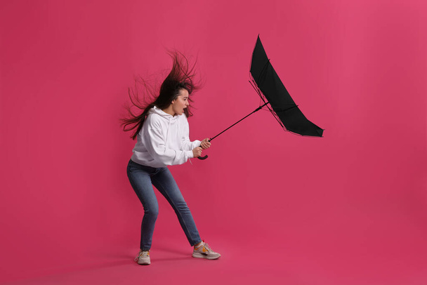 Emotional woman with umbrella caught in gust of wind on pink background - Фото, изображение