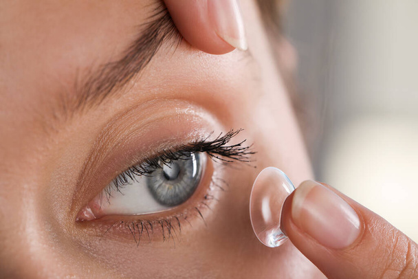 Young woman putting contact lens in her eye on blurred background, closeup - Foto, Bild