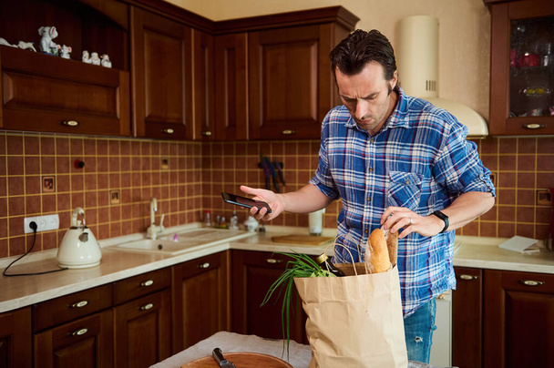 Healthy shopping at grocery store. Food delivery. Handsome middle-aged Caucasian man checking costs while returning home after grocery shopping, sorting organic vegetables and food while unpacking bag - Foto, Bild