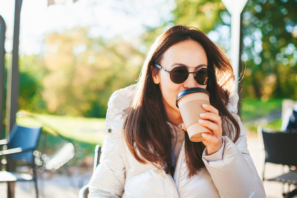 Young woman drinking coffee from disposable coffee cup to go sitting in the sunny outdoors cafe - Foto, immagini