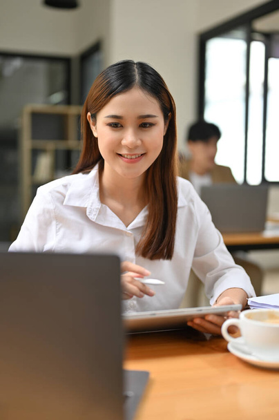 Портрет, красивая и очаровательная молодая азиатская бизнесвумен или работница офиса смотрит на экран своего ноутбука, работая над своим проектом на планшете за своим столом. - Фото, изображение