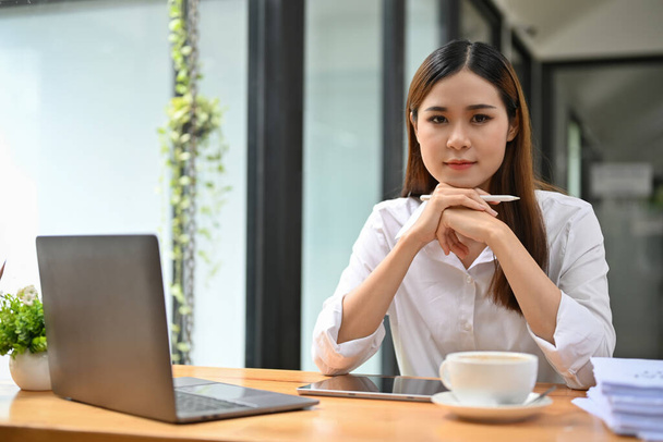 Confident and determined young Asian businesswoman at her office desk hands on chin, smiling and looking at camera. - Foto, Bild