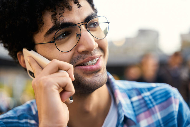 Handsome middle eastern male student calling to friend sitting in cafe. Smiling multiracial hipster guy talking mobile phone outdoors  - Photo, Image