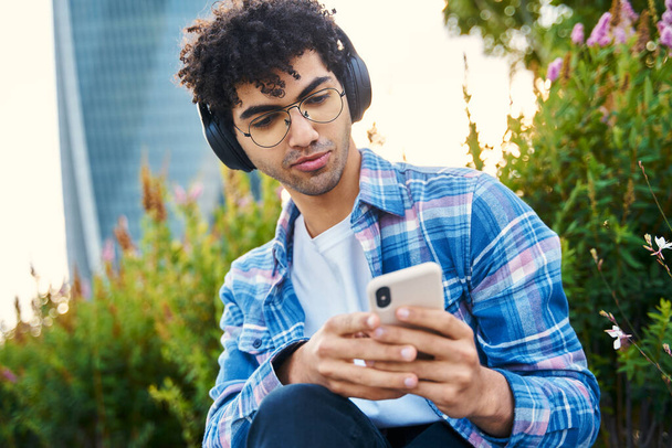Handsome curly haired man wearing headphones listening music, reading text message, communication online outdoors. Pensive middle eastern student holding smartphone sitting in park   - Photo, Image