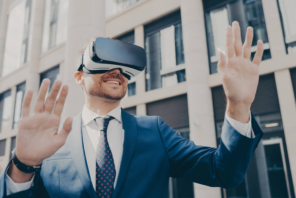 Professioneller aufgeregter Büroangestellter im blauen Anzug, der allein draußen steht und mit einer VR-Brille Projekte visualisiert, etwas in virtueller Realität mit den Händen hält, im Hintergrund baut - Foto, Bild